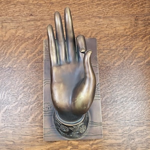 Può includere: Una scultura in metallo color bronzo raffigurante una mano aperta con le dita distese e il pollice che tocca l'indice, montata su una base rettangolare. La base presenta motivi a linee incise e un decoro floreale all'altezza del polso.