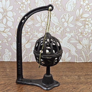 May include: A dark gray cast iron yarn holder featuring a curved stand with a base and a suspended, spherical, open-lattice cage. A thin green string hangs from the top of the curved arm into the center of the cage, which sits on a pedestal.