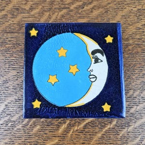 May include: A square ceramic tile with a dark blue crackle glaze finish. The centre features a light blue circular moon with a white crescent profile showing a human face. Five yellow stars are scattered across the blue surfaces.