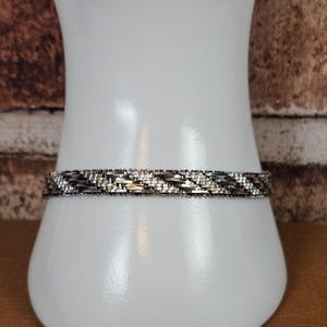 Könnte beinhalten: Ein metallisches Armband mit einem gewebten, mehrfarbigen Design aus abwechselnden silbernen und dunkelgrauen Gliedern. Das Band hat eine texturierte, geflochtene Optik mit kleinen, gleichmäßigen Segmenten in einem sich wiederholenden Muster.
