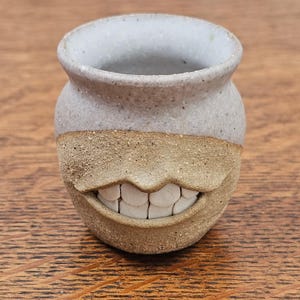 Puede incluir: Una pequeña taza de cerámica con un esmalte gris claro moteado en la parte superior y un acabado de arcilla color canela texturizada en la parte inferior. La taza presenta una boca esculpida con dientes blancos visibles en el centro.