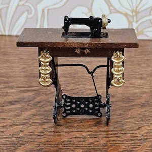Può includere: Una macchina da cucire in miniatura con testina in metallo nero e ruota dorata, montata su un tavolo in legno marrone. La base presenta gambe in metallo nero con decorazioni dorate e un pedale in metallo nero nella parte inferiore.