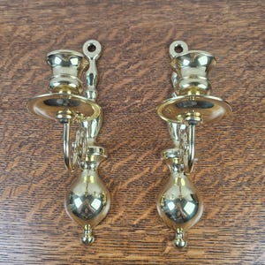 May include: A pair of polished brass wall sconce candle holders featuring a vertical mounting plate with a circular hole at the top, a curved arm, a wide drip tray, and a teardrop-shaped base. The metal surface is reflective and smooth.