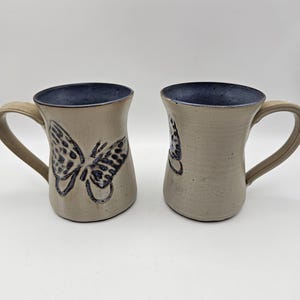 May include: Two ceramic mugs with a butterfly design. The mugs are beige with a dark blue interior and handle. The butterfly design is dark blue. The mugs are handmade and have a rustic appearance.