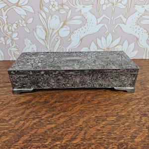 Puede incluir: Una caja rectangular de metal plateado con un intrincado patrón floral y de enredaderas en relieve en la tapa y los laterales. La caja tiene una placa central lisa de forma ovalada en la tapa y se apoya sobre cuatro patas pequeñas y firmes.