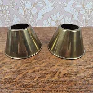 May include: A pair of matching brass candle holders with a smooth, conical shape and a wide base. Each holder features a circular opening at the top for a candle, with a polished metallic finish that reflects light across the surface of the metal.
