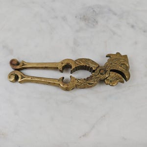 May include: A brass nutcracker shaped like a rooster. The tool features two hinged handles with curved ends and a central circular opening with textured inner edges for gripping. The head of the rooster forms the front end of the metal device.