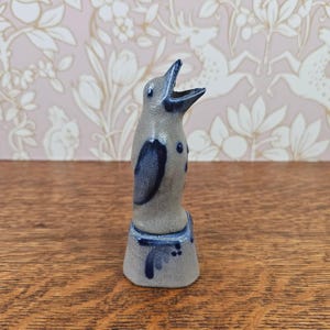 Puede incluir: Una figura de cerámica de un pingüino con el pico abierto, que presenta un cuerpo gris claro moteado y detalles en azul oscuro en las alas, el pecho y la base. El pingüino está de pie sobre una base hexagonal con un acabado esmaltado.