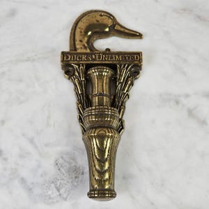 May include: A brass wall-mounted hook featuring a duck head silhouette at the top and the text "DUCKS UNLIMITED" embossed across the center. The lower portion is shaped like a duck call, flanked by decorative reed or feather motifs on both sides.