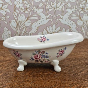 May include: A cream-colored ceramic bathtub-shaped dish featuring four claw feet and a floral pattern of pink roses and blue flowers on the exterior and interior rim. The piece has a smooth, glossy finish and a curved, open-top basin design.