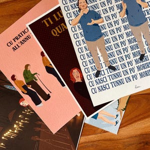 Könnte beinhalten: Eine Sammlung von Kunstdrucken mit Illustrationen und Text. Die Drucke zeigen Figuren und Text in verschiedenen Farben, darunter Rosa, Braun und Weiß. Die Drucke sind auf einer Holzoberfläche angeordnet.