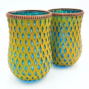 May include: Two glass candle holders featuring a vibrant yellow exterior with a diamond-shaped lattice pattern that reveals a bright blue interior. Each vessel is finished with a textured copper-colored metal rim along the top edge.