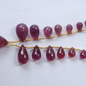 Può includere: Due fili di pietre preziose lucide a forma di goccia di varie dimensioni. Le gemme sono di un profondo colore rosso-viola traslucido, infilate su un sottile filo d'oro. La disposizione suggerisce una collana o un filo di perle.