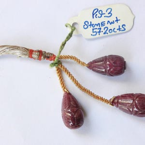 May include: A handcrafted jewelry piece featuring three carved, dark red gemstone pendants suspended from gold-colored twisted cords. The piece is attached to a silver-colored tassel with red accents and a tag that reads "Ps-3 StemeNt 57.20cts".