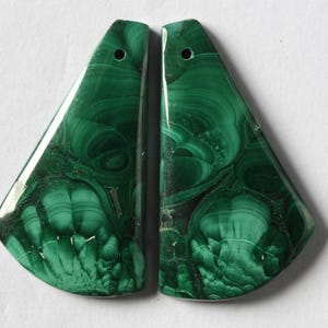 Puede incluir: Dos colgantes de piedra malaquita pulida. Cada colgante tiene forma triangular con un agujero perforado en la parte superior. La piedra verde tiene un patrón arremolinado de verde oscuro y claro.