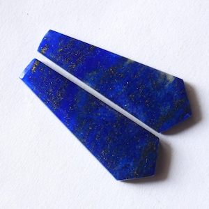 May include: Two polished lapis lazuli gemstone pieces. Each piece is a deep blue color with gold flecks, and a trapezoid shape. The gemstones are displayed on a white surface.