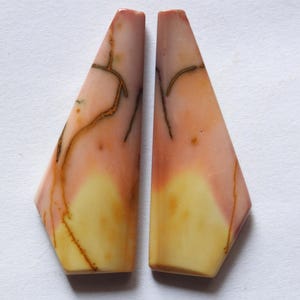 May include: Two polished gemstone pieces. Each piece is a unique shape, with a light pink top fading into a yellow base. Brown vein-like patterns run across the surface. The gemstones are set against a white background.