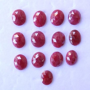 May include: A collection of twelve oval-shaped, faceted, deep red gemstones. The gems are arranged in a grid pattern against a white background, showcasing their polished surfaces and internal clarity. These stones are suitable for jewelry making.
