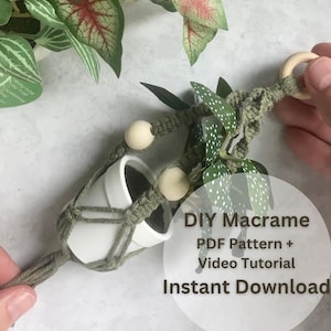 Könnte beinhalten: Ein handgefertigter Makramee-Pflanzenaufhänger in Olivgrün, der zwei kleine weiße Töpfe hält. Das Design umfasst Holzperlen und einen Holzring. Der Text "DIY Makramee PDF Pattern + Video Tutorial Instant Download" ist ebenfalls enthalten.