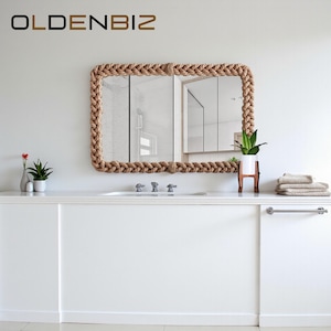 May include: A rectangular mirror with a braided rope frame hangs above a white countertop. The mirror reflects a bathroom interior. The countertop features a sink, plants, and folded towels. The brand name "OLDENBIZ" is in the top left corner.