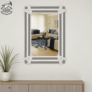 May include: A rectangular mirror with a rope-style frame. The frame features alternating sections of gray and white rope, creating a nautical aesthetic. The mirror reflects a living room with beige furniture and blue accents. The logo "OLDENBIZ" is in the top left corner.