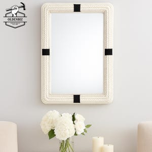 May include: Rectangular wall mirror with a cream-colored rope-style frame. The frame is accented with black bands at each corner. The mirror is hanging on a white wall. The Oldenbiz logo is in the upper left corner.
