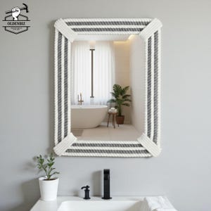 May include: Rectangular mirror with a rope-style frame in white and gray. The mirror reflects a bathroom scene with a white bathtub, plants, and a window. The mirror is a decorative item for the bathroom.