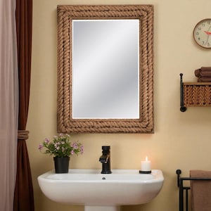 May include: Rectangular mirror with a thick, rope-style frame. The mirror hangs above a white sink with a black faucet. A small potted plant and a lit candle are on the counter. A clock and a shelf with towels are visible in the background.