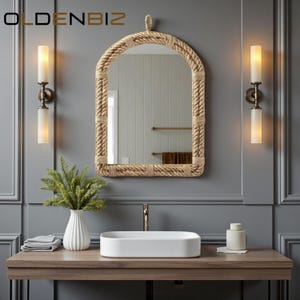 Puede incluir: Un mueble de baño con un espejo enmarcado con cuerda, un lavabo rectangular blanco y dos apliques. El espejo tiene una parte superior arqueada. Un jarrón blanco con vegetación y un tarro pequeño se encuentran sobre la encimera de madera. La pared está pintada de gris.