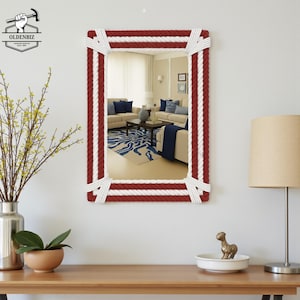 Op de afbeelding: Een rechthoekige spiegel met een nautisch thema. Het frame is gemaakt van rood en wit touw, met witte touwaccenten op de hoeken. De spiegel weerspiegelt een woonkamer met een bank, vloerkleed en lamp.