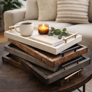 May include: A stack of wooden serving trays in various shades of brown and gray, with gold and black handles. The top tray holds a white mug, a lit candle, a book with the word "STILLNESS", and greenery. The trays are on a dark wooden table.