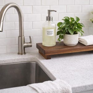 Pode incluir: Uma bancada de cozinha com uma torneira e uma pia de a&ccedil;o inoxid&aacute;vel. Uma bandeja de madeira cont&eacute;m um dispensador de sabonete l&iacute;quido transparente, uma pequena planta em vaso e uma toalha branca dobrada. O dispensador de sabonete tem o r&oacute;tulo "HAND SOAP".