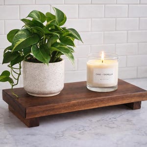 May include: A wooden decorative tray holds a potted plant and a lit candle. The plant has green and yellow leaves in a speckled white pot. The candle is in a clear glass with a label that reads "Oak - Vanilla".