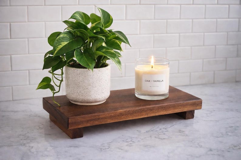 Pode incluir: Uma bandeja decorativa de madeira escura exibe uma planta em vaso e uma vela acesa. A planta tem folhas verdes e amarelas num vaso branco salpicado. A vela est&aacute; num copo transparente com o r&oacute;tulo "Oak - Vanilla".