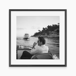 May include: Black and white photograph of a person on a boat, holding a glass of a beverage. The person is wearing sunglasses and a button-down shirt. A boat is in the background, with land visible on the horizon.