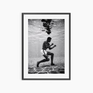 Puede incluir: Impresión enmarcada en blanco y negro de un boxeador bajo el agua, en posición de combate. El boxeador lleva pantalones cortos blancos con ribete negro. La superficie del agua y el reflejo del boxeador son visibles.