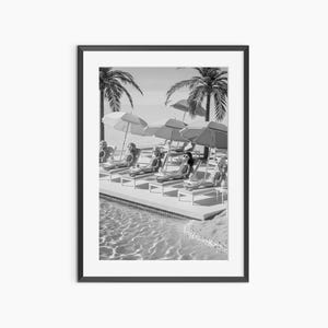 May include: Black and white framed print of dolls lounging by a pool under umbrellas. Palm trees and a sandy beach are in the background. The dolls are wearing sunglasses and swimsuits, relaxing on white lounge chairs.