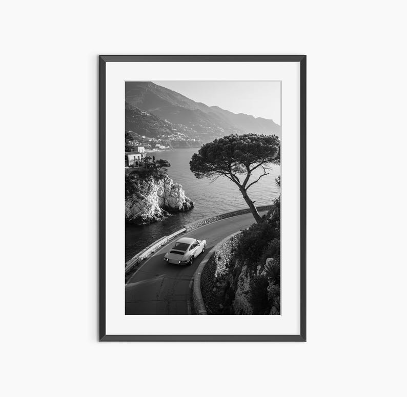 A black and white photograph of a car driving along a winding coastal road, with a large tree on the side of the road and mountains visible in the background.