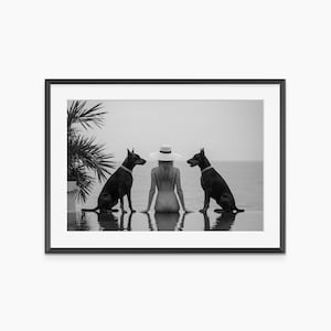 May include: Black and white framed print featuring two black dogs flanking a person wearing a white hat, seated near water. A palm tree is visible on the left. The background shows the ocean and sky.