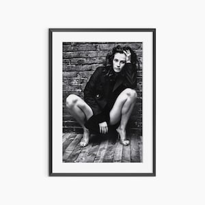 Può includere: Fotografia incorniciata in bianco e nero di una persona accovacciata, con un cappotto scuro e tacchi alti. Lo sfondo presenta un muro di mattoni e assi di legno. La fotografia è un ritratto.