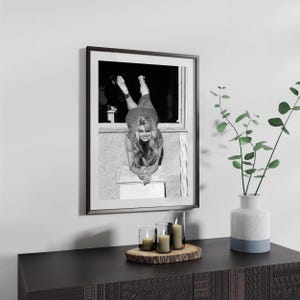 a framed black and white photograph of a woman leaning on a window sill, with a vase of greenery and candles on a wooden surface below the frame.