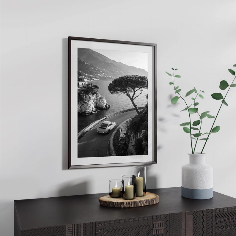 a black and white photograph of a winding road with a car driving along it, framed and hanging on a wall. Below the frame, there is a wooden table with a vase of greenery and three candles.