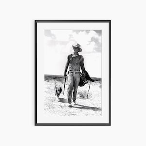 May include: Black and white framed print of a cowboy walking across a desert landscape, holding a riding crop and a saddle. A dog walks alongside him. The cowboy wears a hat, shirt, jeans, and boots.