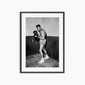 Puede incluir: Fotografía en blanco y negro enmarcada de un boxeador en posición de combate. El boxeador lleva guantes negros, pantalones cortos y calcetines y zapatos blancos. El fondo es una pared y un suelo de madera.