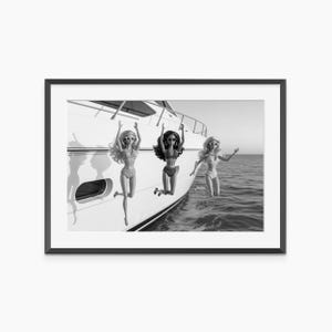 May include: Black and white framed print featuring three dolls in swimwear, posing near a yacht. The dolls are wearing sunglasses and have their arms raised. The image is set against a backdrop of the ocean and sky.