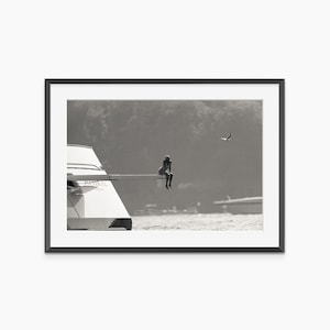 Może przedstawiać: Czarno-biała fotografia osoby siedzącej na krawędzi jachtu, z ptakiem w tle. Jacht jest biały, a woda i niebo są w delikatnej szarości. Czarna ramka.