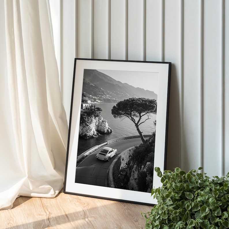 A framed black and white photograph of a car driving on a winding road near a body of water, with a tree and mountains in the background.