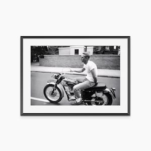 May include: Black and white framed print of a man on a vintage motorcycle. The motorcycle has a silver gas tank, black seat, and chrome handlebars. The man is wearing a white t-shirt and jeans. The license plate reads DN18228.