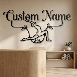 May include: A black metal wall sign featuring the text Custom Name in a cursive script font, positioned above a silhouette of a person in a yoga pose, flanked by two leaf stems and a lotus flower icon centered at the bottom of the design.