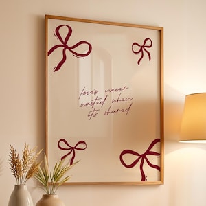 May include: A framed print with a cream background and burgundy bow accents. The print features the quote "Love's never wasted when it's shared." A wooden lamp and two vases with plants sit on a wooden shelf.